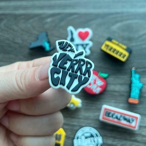May include: A collection of colorful shoe charms featuring New York City themes. Includes a black and white apple with "VERRR CITY" text, an "I Love NY" charm, a yellow taxi, and a miniature Statue of Liberty.