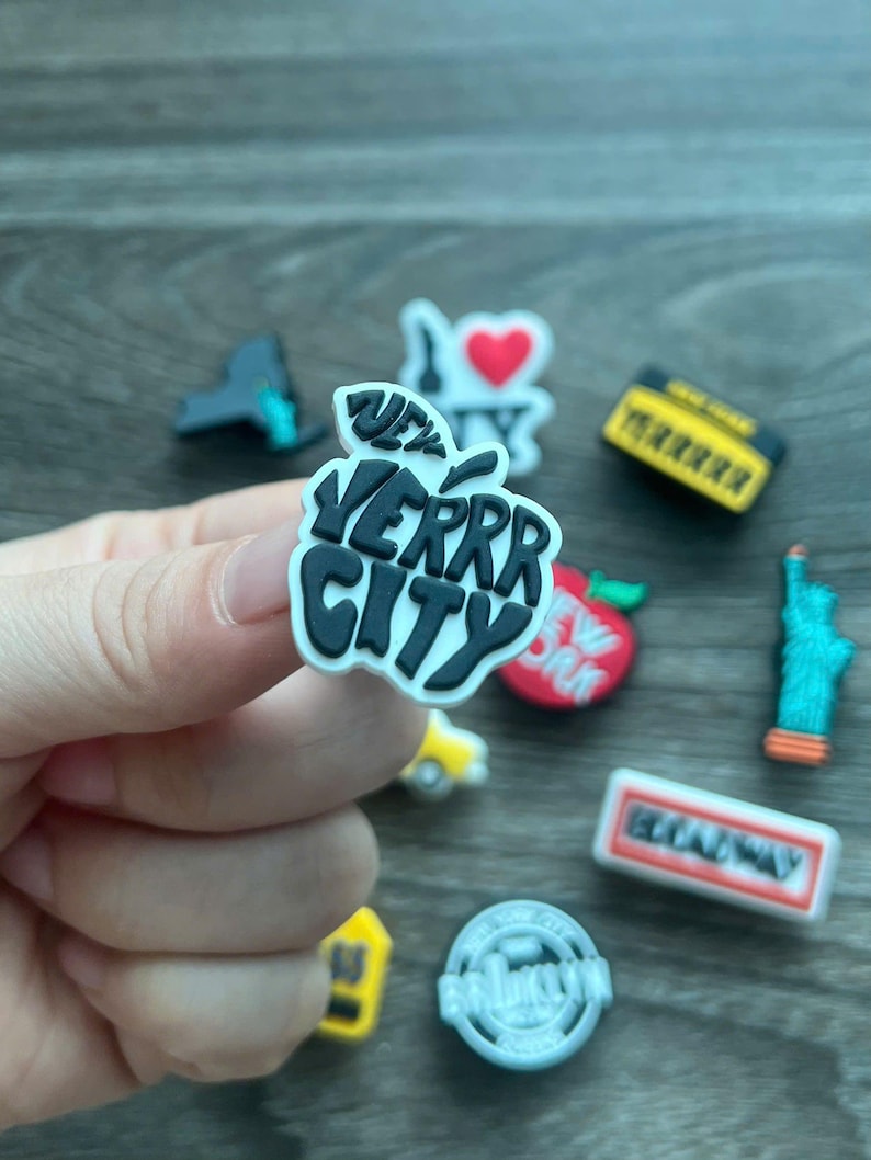 May include: A collection of colorful shoe charms featuring New York City themes. Includes a black and white apple with "VERRR CITY" text, an "I Love NY" charm, a yellow taxi, and a miniature Statue of Liberty.