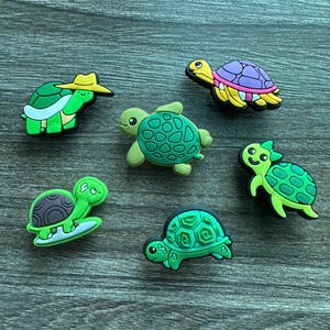 May include: A collection of colorful turtle-shaped charms. The charms feature various designs, including a turtle wearing a hat, a turtle with a purple shell, and a turtle on a surfboard. The charms are in shades of green, brown, and yellow.