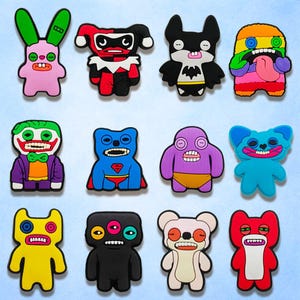 May include: A collection of colorful cartoon character-shaped rubber charms. The charms feature various designs, including a pink bunny, a red and black jester, a black bat, and a rainbow-colored monster. Other characters include a Joker, Superman, and a purple creature.