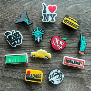 May include: A collection of New York City-themed shoe charms. The charms feature iconic landmarks like the Statue of Liberty, a yellow taxi, and the New York skyline. Other charms display the words "NY", "Broadway", and "Wall St".