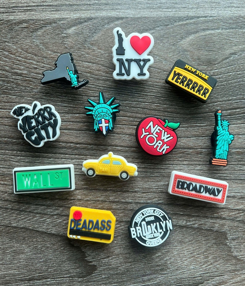 May include: A collection of New York City-themed shoe charms. The charms feature iconic landmarks like the Statue of Liberty, a yellow taxi, and the New York skyline. Other charms display the words "NY", "Broadway", and "Wall St".