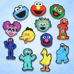 May include: A collection of colorful Sesame Street character charms, including Cookie Monster, Elmo, Big Bird, and others. These flat, cartoon-style charms are in various colors like blue, red, yellow, and green, perfect for crafting or personalizing accessories.