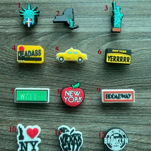 May include: A collection of New York City-themed shoe charms. Designs include the Statue of Liberty, a yellow taxi, the New York skyline, and the words "DEADASS", "YERRRRR", "WALL ST", "NEW YORK", "BROADWAY", "NY", "VERRR CITY", and "BROOKLYN".