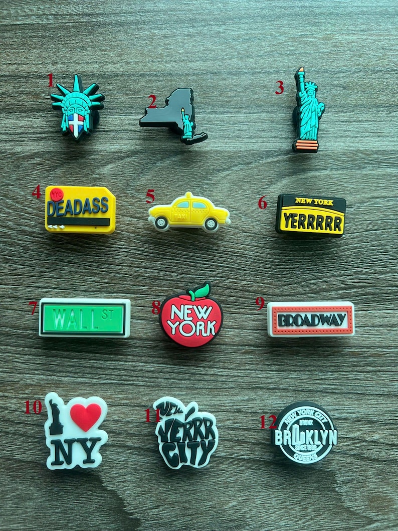 May include: A collection of New York City-themed shoe charms. Designs include the Statue of Liberty, a yellow taxi, the New York skyline, and the words "DEADASS", "YERRRRR", "WALL ST", "NEW YORK", "BROADWAY", "NY", "VERRR CITY", and "BROOKLYN".