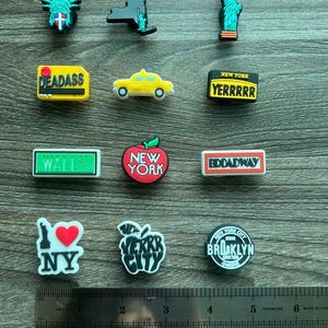May include: A collection of New York City-themed charms. The charms include the Statue of Liberty, the New York skyline, a yellow taxi, and signs that read "DEADASS", "YERRRRR", "WALL ST", and "BROADWAY". Also included are charms with an apple and the text "NY", "VERKR CITY", and "BROOKLYN QUEENS".