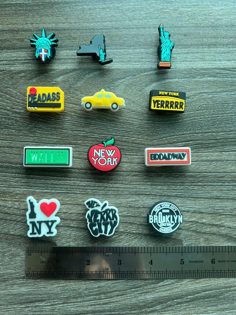 May include: A collection of New York City-themed charms. The charms include the Statue of Liberty, the New York skyline, a yellow taxi, and signs that read "DEADASS", "YERRRRR", "WALL ST", and "BROADWAY". Also included are charms with an apple and the text "NY", "VERKR CITY", and "BROOKLYN QUEENS".