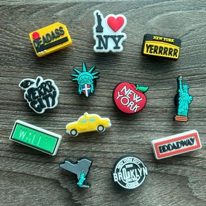 May include: A collection of colorful New York City-themed shoe charms. Designs include the Statue of Liberty, a yellow taxi, an apple, and various text-based charms with phrases like "NY", "Broadway", and "Wall St."