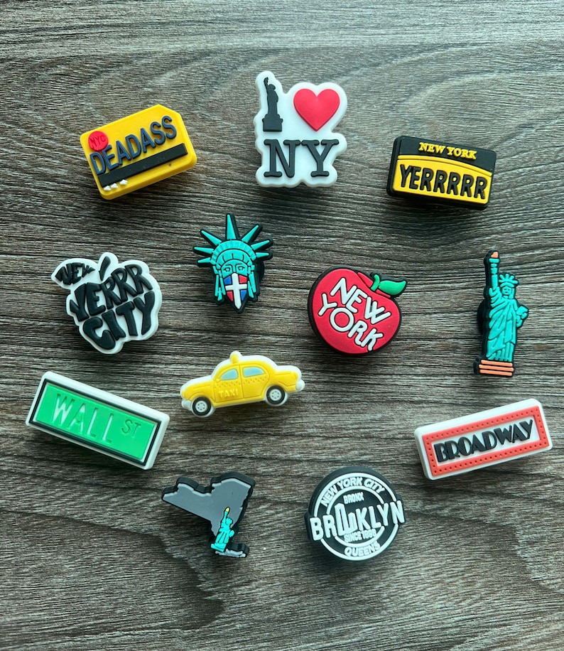 May include: A collection of colorful New York City-themed shoe charms. Designs include the Statue of Liberty, a yellow taxi, an apple, and various text-based charms with phrases like "NY", "Broadway", and "Wall St."