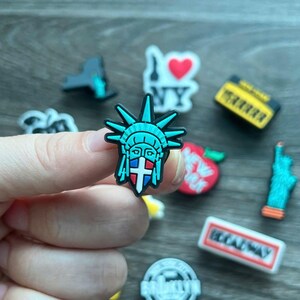 May include: A collection of colorful shoe charms featuring iconic New York City landmarks. The charms include the Statue of Liberty, the NY heart logo, a yellow taxi, a red apple, and a Broadway sign. The Statue of Liberty charm is held by a hand.