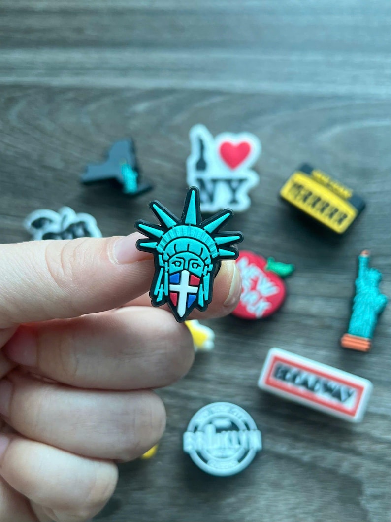 May include: A collection of colorful shoe charms featuring iconic New York City landmarks. The charms include the Statue of Liberty, the NY heart logo, a yellow taxi, a red apple, and a Broadway sign. The Statue of Liberty charm is held by a hand.