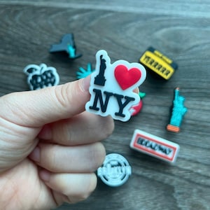 May include: A collection of colorful shoe charms featuring iconic New York City landmarks. The charms include the Statue of Liberty, an apple, a taxi cab, and a charm that says "I Love NY" with a red heart.