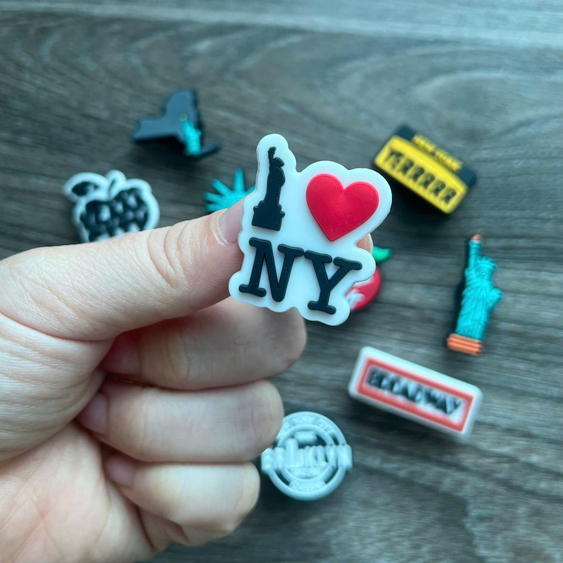 May include: A collection of colorful shoe charms featuring iconic New York City landmarks. The charms include the Statue of Liberty, an apple, a taxi cab, and a charm that says "I Love NY" with a red heart.
