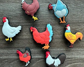 May include: A collection of seven colorful chicken-shaped charms. The charms feature various colors, including red, white, blue, and yellow, with black outlines. Each charm has a unique design, showcasing different chicken poses and details.