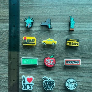 May include: A collection of New York City-themed charms, including the Statue of Liberty, a yellow taxi, and an apple with "New York" text. Other charms feature the text "DEADASS", "YERRRRR", "WALL ST", "BROADWAY", "I ❤️ NY", and "BROOKLYN QUEENS".