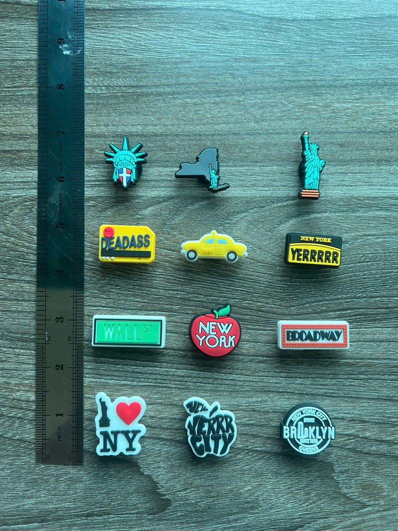 May include: A collection of New York City-themed charms, including the Statue of Liberty, a yellow taxi, and an apple with "New York" text. Other charms feature the text "DEADASS", "YERRRRR", "WALL ST", "BROADWAY", "I ❤️ NY", and "BROOKLYN QUEENS".