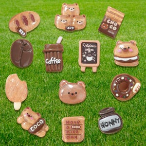 May include: A collection of brown and tan food-themed charms, including a coffee cup, bear-shaped pastries, a chocolate bar, and a donut. The charms are arranged on a green grassy surface.