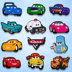 May include: A collection of colorful car-shaped charms, featuring various designs and characters. The charms include a blue race car, a green sports car, and a purple hot rod. The word "Cars" is displayed on a black and red sign.