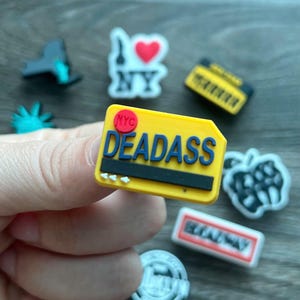 May include: A collection of colorful shoe charms. One charm is a yellow rectangle with the word "DEADASS" in blue, and "NYC" in red. Other charms include a yellow boombox, a Statue of Liberty, and an "I Love NY" design.