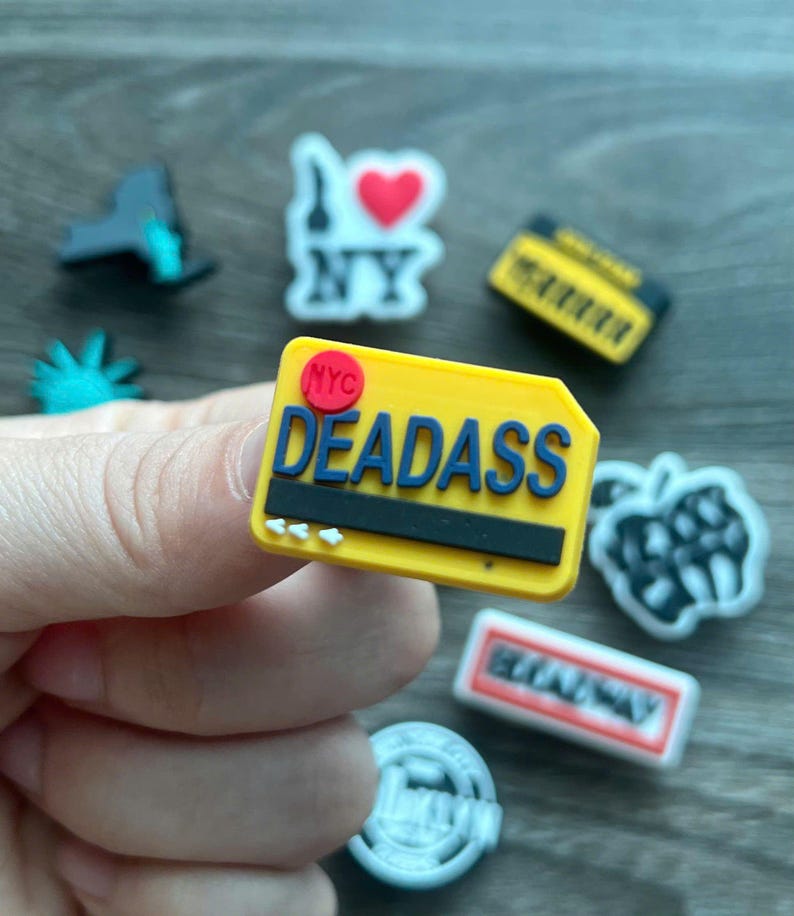 May include: A collection of colorful shoe charms. One charm is a yellow rectangle with the word "DEADASS" in blue, and "NYC" in red. Other charms include a yellow boombox, a Statue of Liberty, and an "I Love NY" design.