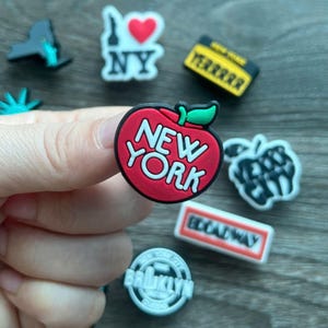May include: A collection of New York City-themed charms. The charms include a red apple with "NEW YORK" text, a yellow taxi cab, a "Broadway" sign, and a "Brooklyn" emblem. Other charms feature the Statue of Liberty and an "I Love NY" design.