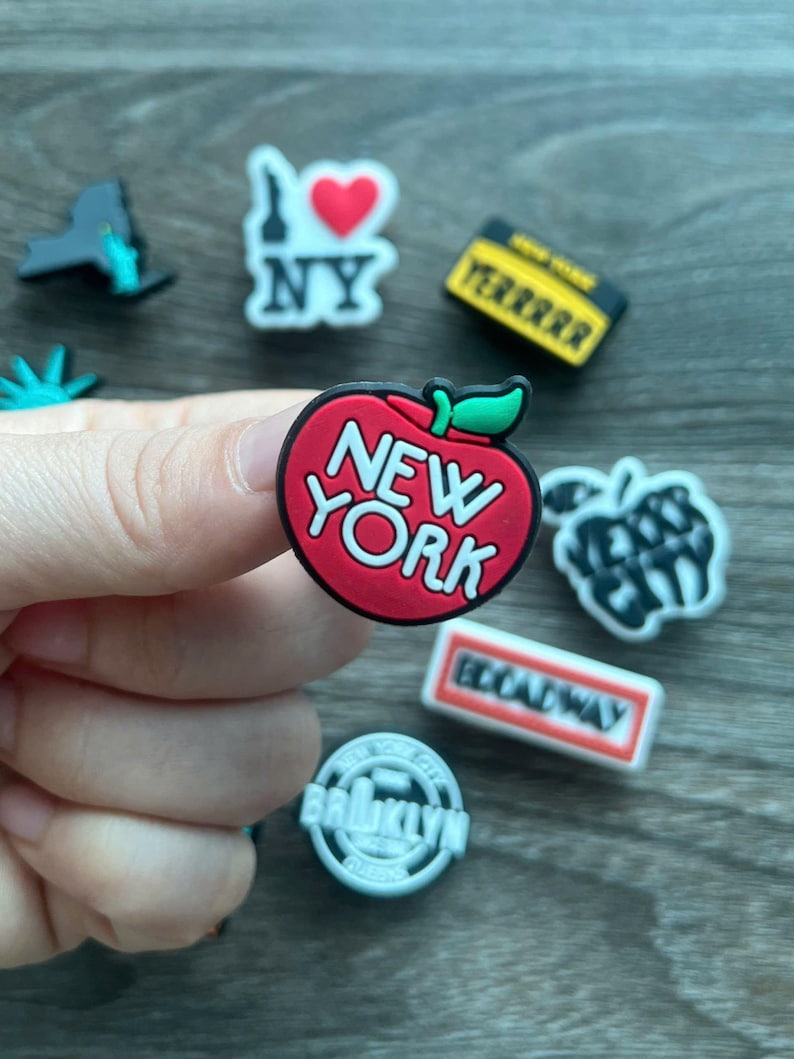 May include: A collection of New York City-themed charms. The charms include a red apple with "NEW YORK" text, a yellow taxi cab, a "Broadway" sign, and a "Brooklyn" emblem. Other charms feature the Statue of Liberty and an "I Love NY" design.