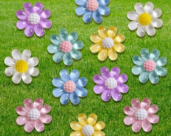 May include: A collection of colorful, translucent flower-shaped embellishments in various shades of pink, purple, blue, and yellow. Each flower has a textured center and is arranged on a green, grassy surface.