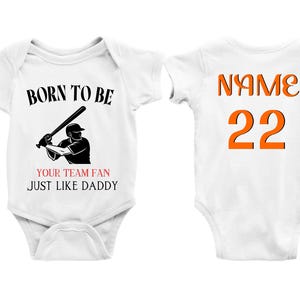 May include: White baby bodysuit featuring the text "BORN TO BE" and a baseball player graphic. Below the graphic, the text reads "YOUR TEAM FAN JUST LIKE DADDY". The back of the bodysuit has the word "NAME" and the number "22" in orange.