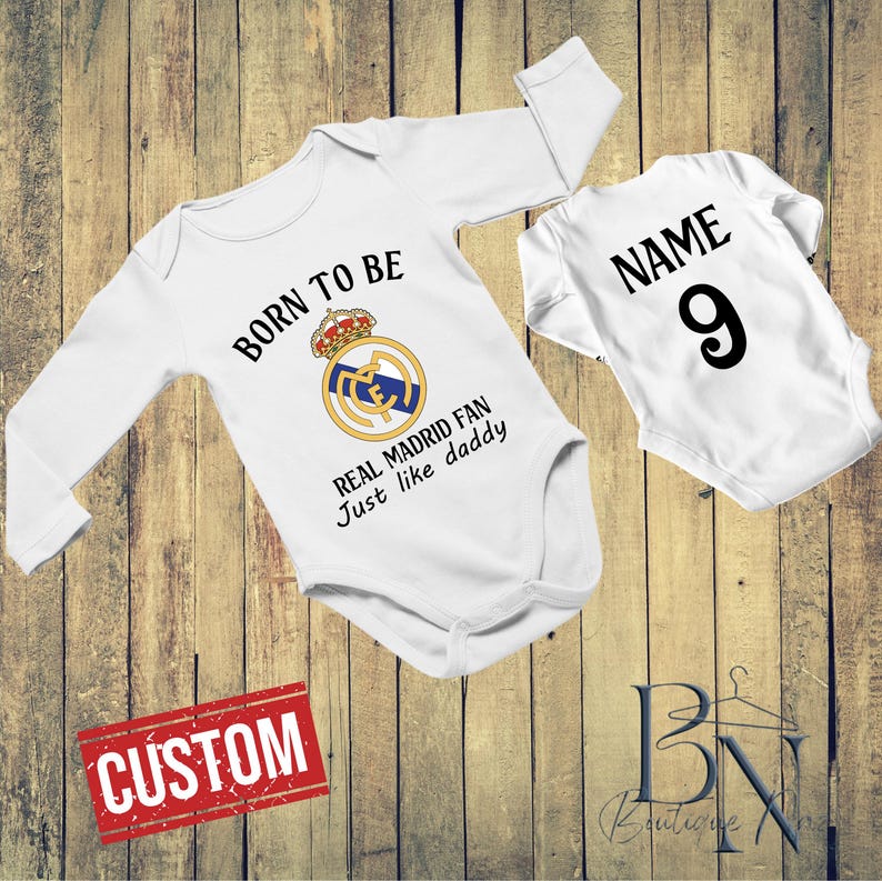 May include: Two white baby onesies with black text and a Real Madrid logo. One says "BORN TO BE REAL MADRID FAN Just like daddy" and the other says "NAME 9". A red "CUSTOM" sign is also visible.