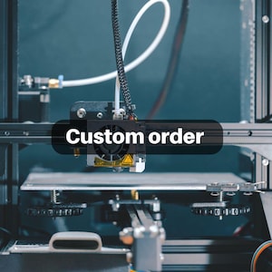 Op de afbeelding: Een 3D-printer met de tekst "Custom order" over het beeld. De printer heeft een zwart frame en een wit printbed. De printkop is zwart met een geel waarschuwingssymbool. Een witte kabel is zichtbaar.
