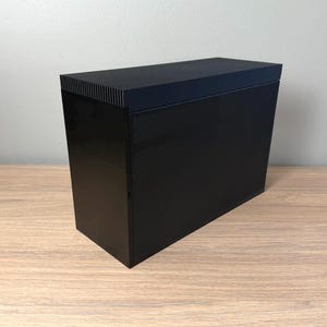 May include: A black rectangular computer case with a textured front panel and a solid top. The case is sitting on a wooden surface. The top of the case has a series of vertical lines.