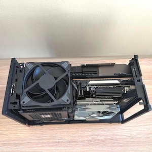 Op de afbeelding: Een zwarte computerkast met zichtbare interne componenten. Een grote zwarte ventilator is links gemonteerd, met een kleinere ventilator rechts. Verschillende printplaten en bedrading zijn ook zichtbaar in de behuizing.