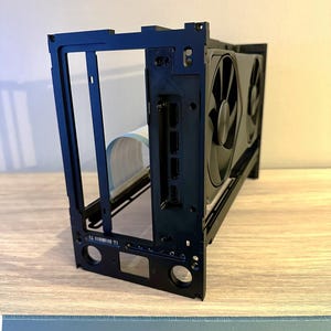 May include: A black computer case frame with a graphics card installed. The frame has a rectangular shape with multiple openings and mounting holes. The graphics card has two black fans.