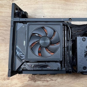 May include: A black computer case with internal components visible. A large black fan with orange accents is centered, with the ROG logo visible on a component below. Wires and other components are also visible.