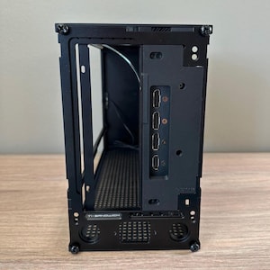 May include: A black computer case with an open frame design. The case has a rectangular shape with visible internal components and a series of ports on the side. The bottom of the case has the text "TI-SANDWICH".