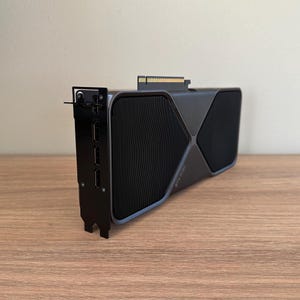 Könnte beinhalten: Eine schwarze und silberne RTX 3090 Grafikkarte mit einem markanten X-förmigen Design und mehreren Anschlüssen. Die Karte hat einen schwarzen Lüfter und der Text "RTX 3090" ist sichtbar.