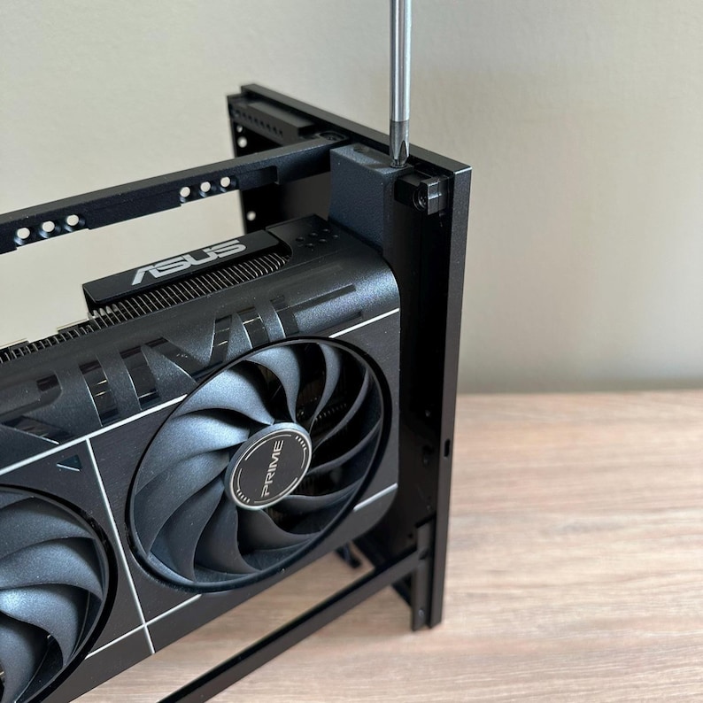 May include: A black ASUS PRIME graphics card is being installed with a screwdriver. The card has two large fans and the ASUS logo. The components are set in a black metal frame.
