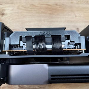 Puede incluir: Vista de cerca de un componente de computadora con el texto "ASKR" visible. El componente es gris y negro, con varios cables negros y una placa de circuito. El dispositivo está montado dentro de una carcasa negra.