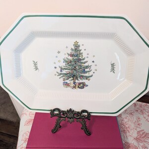 May include: A white octagonal ceramic serving platter with a green rim and a central illustration of a decorated Christmas tree surrounded by small stars and gifts. The platter features a ribbed texture along the inner edges and small sprigs.