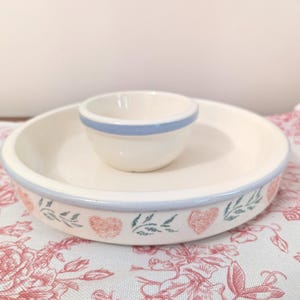 May include: A cream-colored ceramic chip and dip set. The outer bowl has a light blue rim and a decorative border of pink hearts and green leaves. The smaller dip bowl has a light blue rim. The set is on a floral patterned cloth.