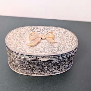 May include: An oval-shaped silver-toned metal trinket box featuring an intricate embossed floral scrollwork pattern across the lid and sides. A prominent, textured gold-toned metal bow is centered on the top of the lid as a decorative accent.