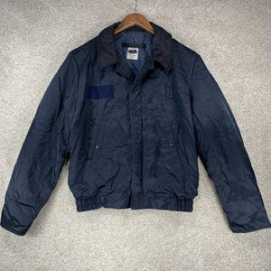 Puede incluir: Chaqueta bomber azul marino con cuello azul oscuro y parche en el pecho izquierdo. La chaqueta tiene cierre de botones en la parte delantera, dos bolsillos delanteros y dobladillo acanalado. La chaqueta está colgada en una percha de madera.