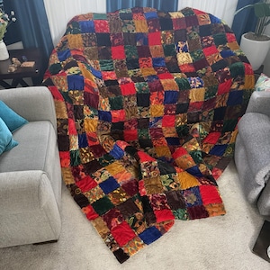 May include: A patchwork quilt with a vibrant mix of colors including red, blue, green, and gold. The quilt is made of square fabric pieces, creating a textured, patterned design. It is draped over a gray sofa, showcasing its size and detail.
