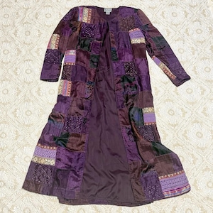 May include: A long-sleeved, open-front duster coat in shades of purple, featuring a patchwork design of various fabric textures, including quilted squares, embroidered panels, and solid satin-like sections, with a solid dark purple interior lining.