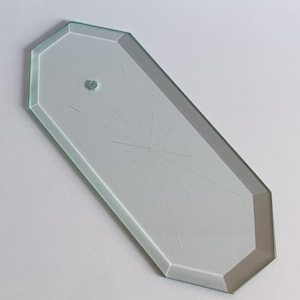 May include: An octagonal clear glass vanity tray with beveled edges. The surface features a delicate, etched starburst pattern radiating from a central point and includes a small, circular glass knob or handle attached near the top edge.