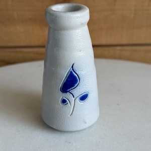 May include: A small, off-white ceramic vase with a blue floral design. The vase has a textured surface and a flared top. The blue flower is stylized and adds a pop of color to the neutral background. The vase is likely for decorative purposes.