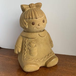 May include: Tan ceramic doll figurine with a bow in her hair and a smiling face. The doll is wearing a dress with a floral design on the front. The figurine is sitting down.