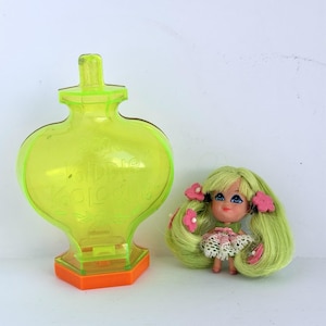 May include: A translucent neon yellow plastic Kiddle Kologne perfume bottle container with an orange base, paired with a small doll featuring long light green hair, pink felt flower hair accessories, and a white lace-trimmed dress.