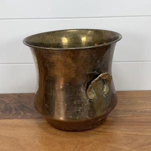 May include: A brass bucket with a wide, flared rim and a tapered base. The metal surface shows a weathered, aged patina with visible dark spots and oxidation. Two ornate, curved handles are attached to the sides of the vessel.