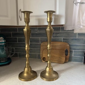 May include: A pair of tall, polished brass candlesticks featuring a flared top, a multi-faceted hexagonal stem, and a wide, circular base. The metallic surface reflects light, showcasing a smooth, uniform finish across the vertical structure.